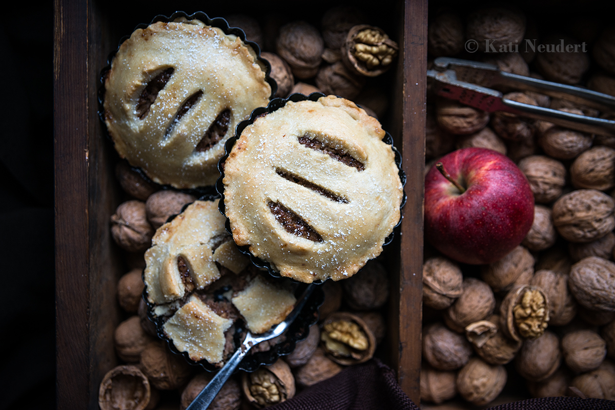 Drei vegane Apfel-Walnuss-Tartelettes liegen zusammen mit Walnüssen und Äpfeln in einer Holzkiste.