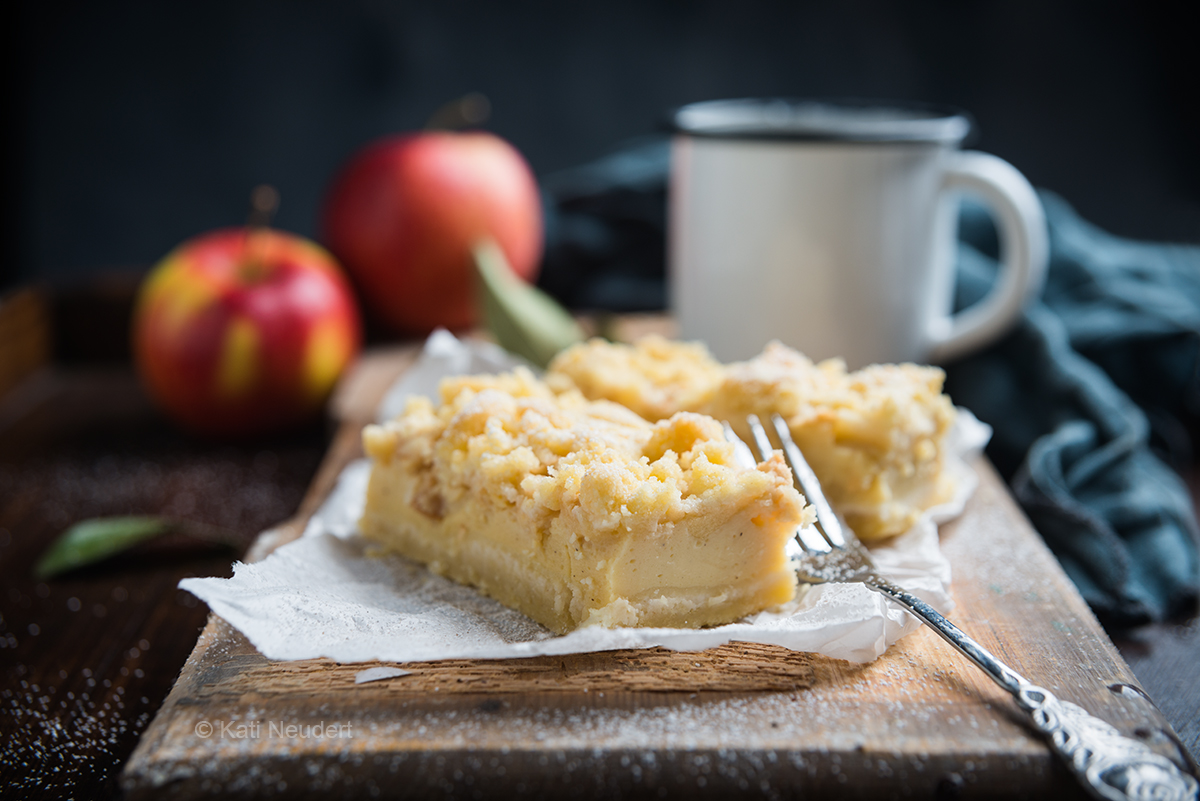 Zwei Stück veganer Apfel-Streusel-Cheesecake auf einem Holztisch zusammen mit Äpfeln und einer Tasse Kaffee.