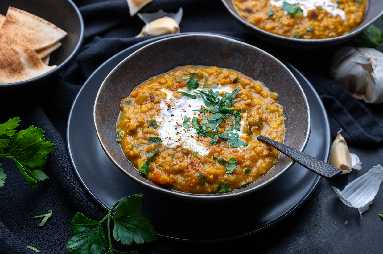 Eine Schüssel mit veganem Dal aus roten Linsen, Tomaten und Kokosmilch. Daneben Fladenbrot, Knoblauch, Petersilie und eine weitere Schale mit Linsen Dal.