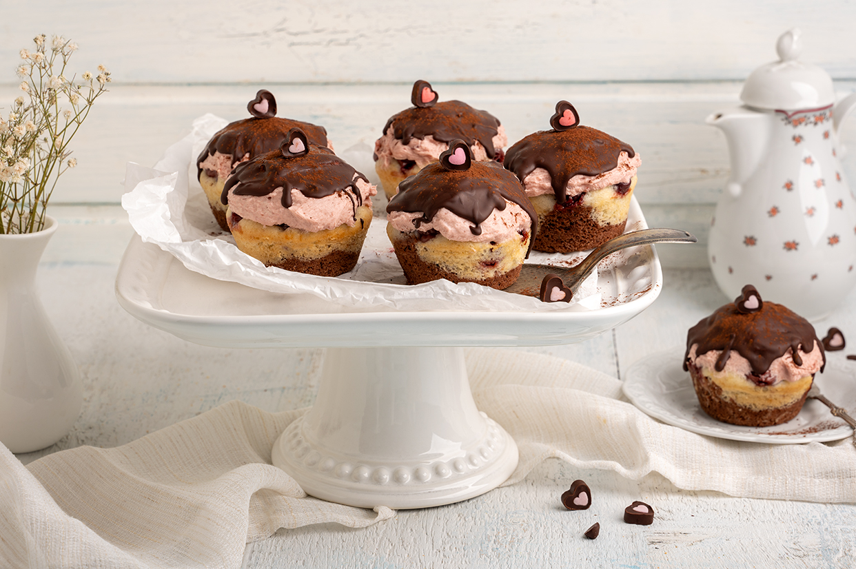 Fünf vegane Cupcakes mit zweierlei Rührteig, Sauerkirschen, Sauerkirsch-Creme und Zartbitterglasur stehen auf einer Tortenplatte. Auf einem Teller daneben steht ein weiterer.