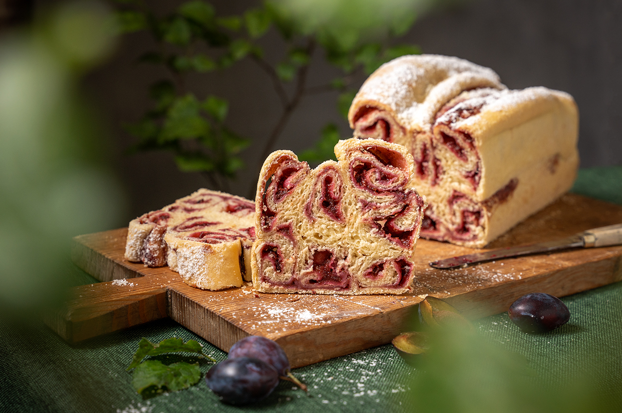 Ein aufgeschnittener Hefekuchen mit Swirlen aus Zwetschgenmus/Zwetschgenmarmelade auf einem Holzbrett,Rundherum frische Zwetschgen und Blätter vom Zwetschgenbaum.