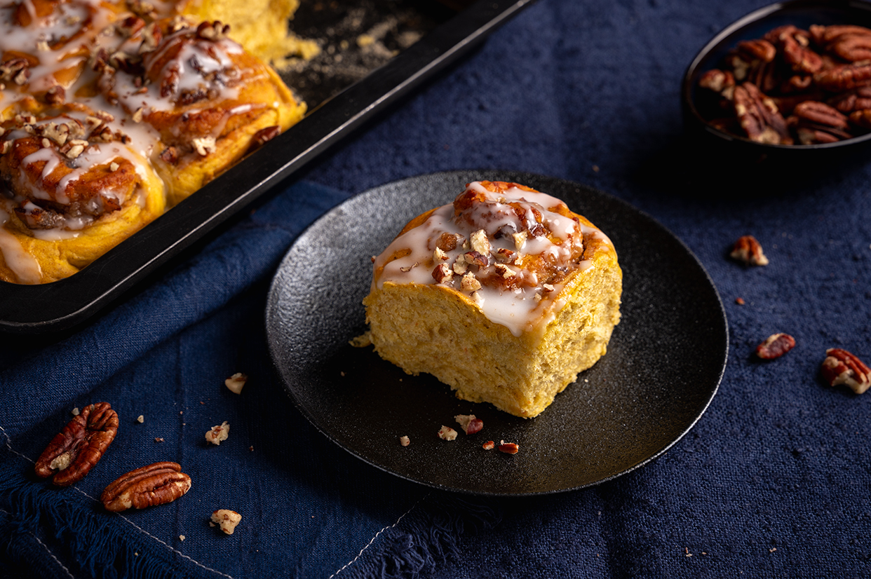 Ein Stück veganer Hefeschnecken-Kuchen mit Kürbis, Mandeln, Pekannüssen und einer Zuckerglasur auf einem schwarzen Teller. Dieser steht auf einem dunkelblauen Untergrund. Links hinten ein Obstkuchenblech mit dem angeschnittenen Kuchen. Rechts hinten eine kleine Schale mit Pekannüssen.