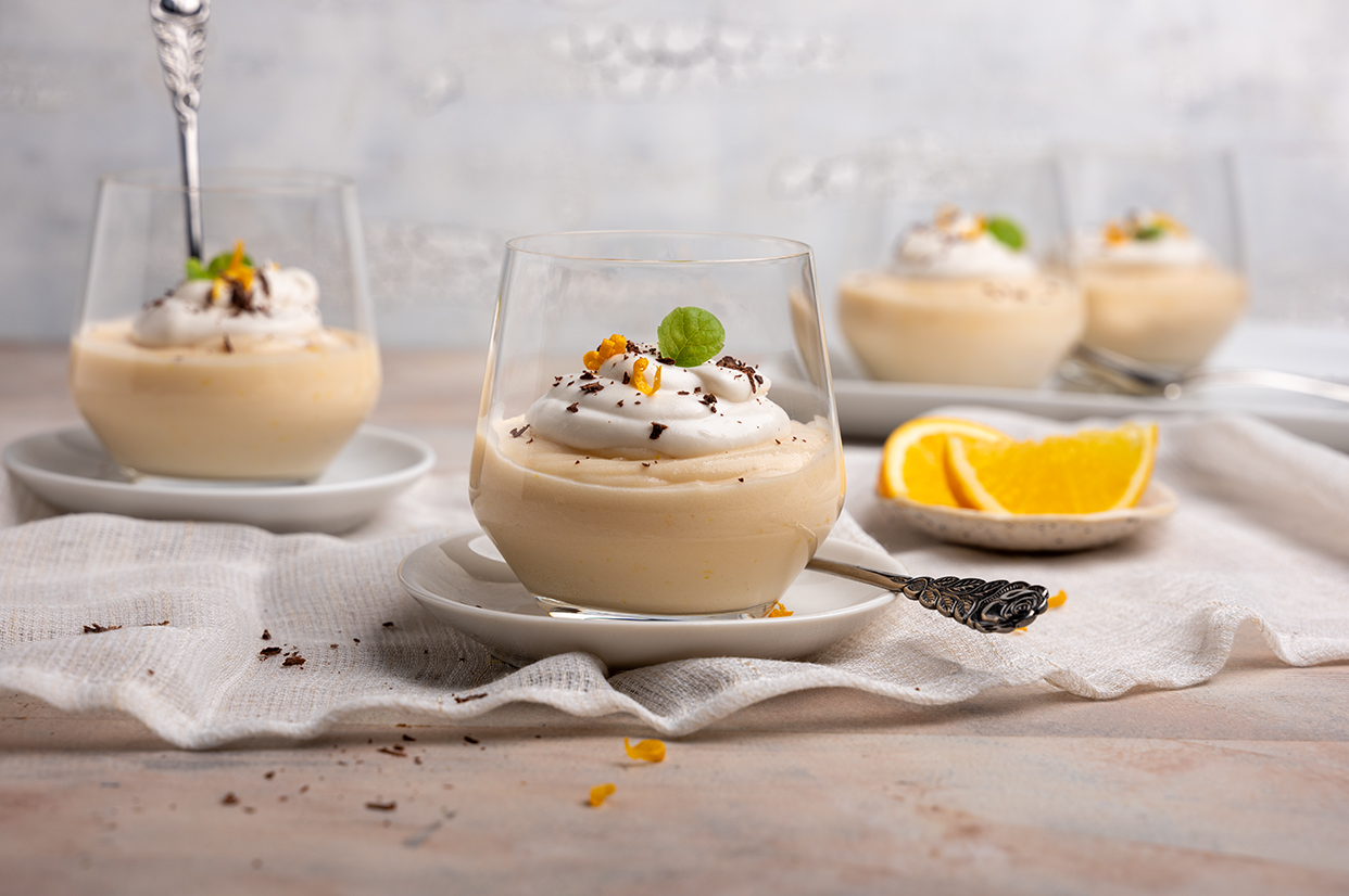 Ein Dessertglas vegane Orangencreme mit Sahnehäubchen, Schokosplittern. Orangenzesten und Minzblättchen im Vordergrund. Drei Gläser mit Dessert und ein Schälchen mit Orangenspalten im Hintergrund. Helles Ambiente.