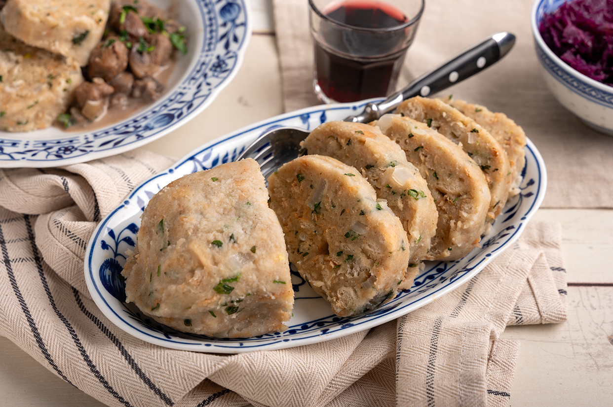 Vegane Serviettenknödel (Semmelknödel) aufgeschnitten im Vordergrund. Im Hintergrund Rotkohl und ein Teller mit Knödeln und veganem Gulasch.
